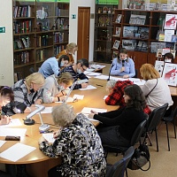 На фото: В читальном зале, за столом сидят читатели и пишут диктант.