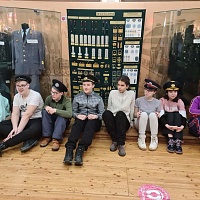 Школьники - участники экскурсии сидят на фоне выставки образцов форменной одежды железнодорожников и знаков отличия в выставочном центре истории Западно-Сибирской железной дороги